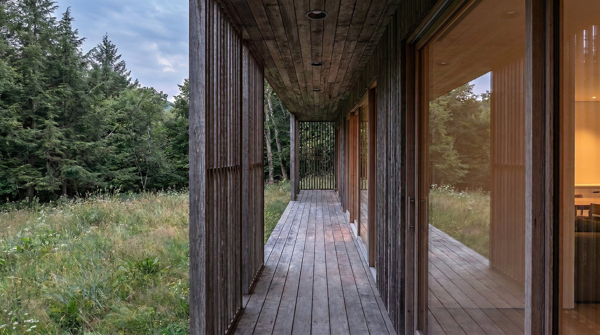 A long wooden deck corridor attached to a modern house, with tall dark wood siding, large glass doors, and an outdoor grassy field and forest visible on both sides.