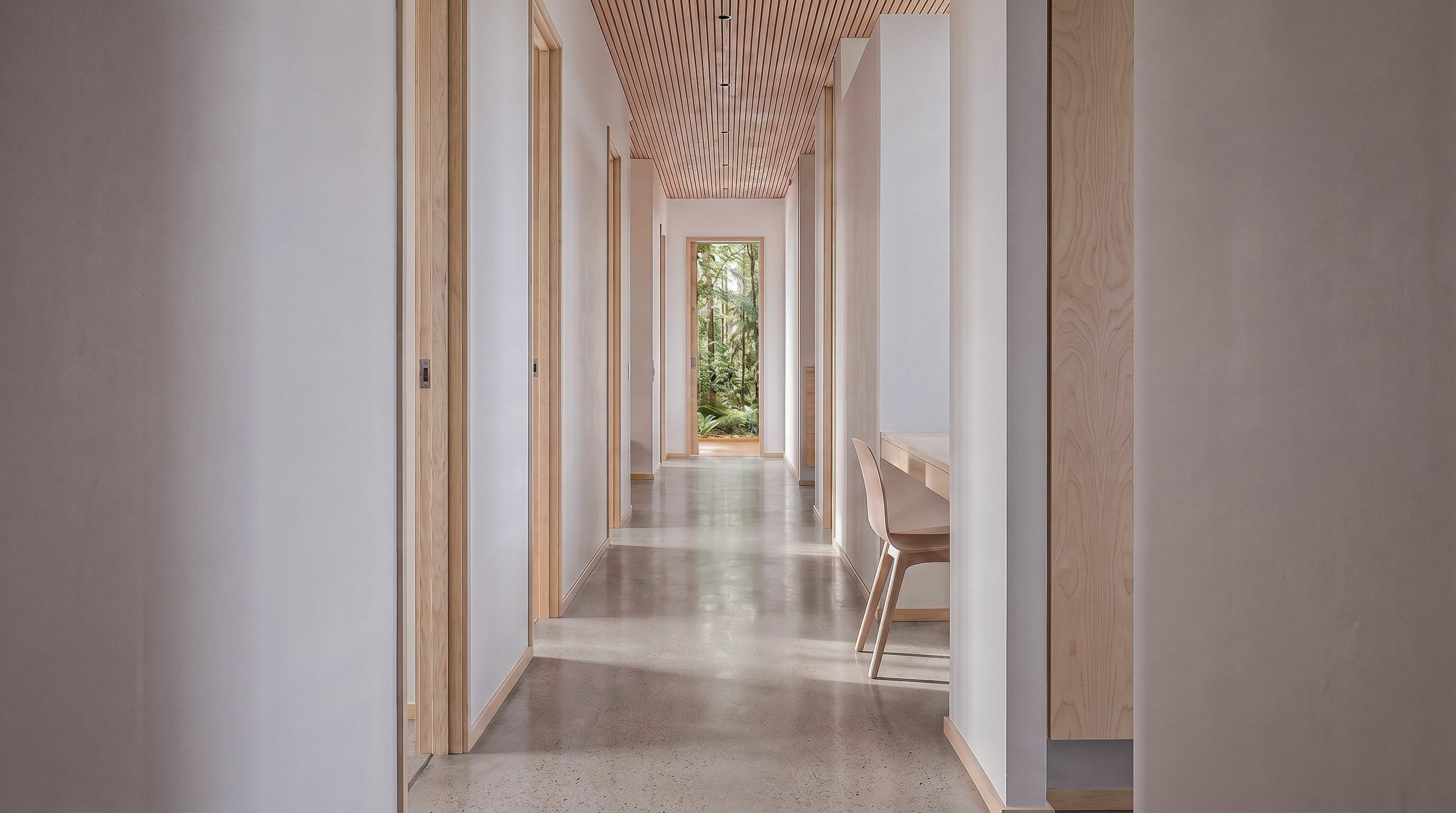 A long, modern hallway with light wood doors, a slatted wood ceiling, and a polished concrete floor, leading to an open window with greenery outside.