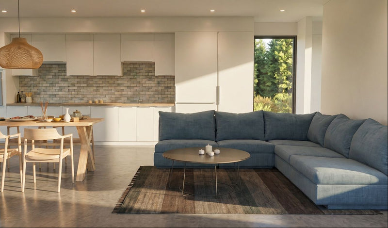 A modern living room featuring a large L-shaped sectional sofa in slate blue, styled with a round wooden coffee table and a textured area rug. In the background, an open-plan kitchen with light wood cabinets and a tiled backsplash is visible, along with a dining table and chairs under a woven pendant light.